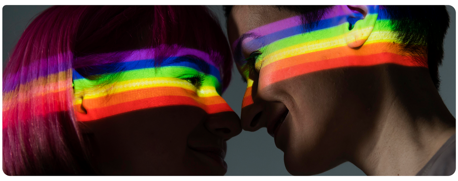 Casal de mulheres se olhando e bem próximas com um arco-íris refletindo nos rostos delas